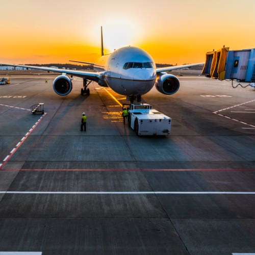 An airliner at its stand on the apron in low sunlight, with a tug and ground crew in front