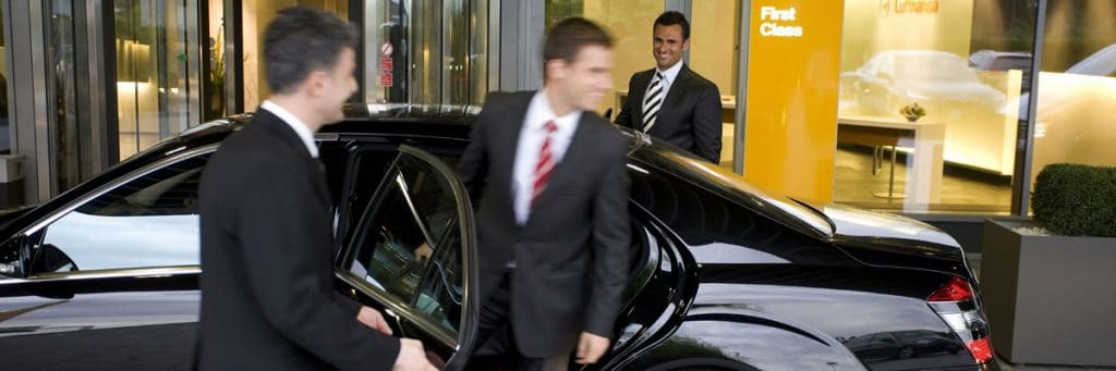 Two men in suits beside a black Mercedes with its rear door open outside a hotel entrance