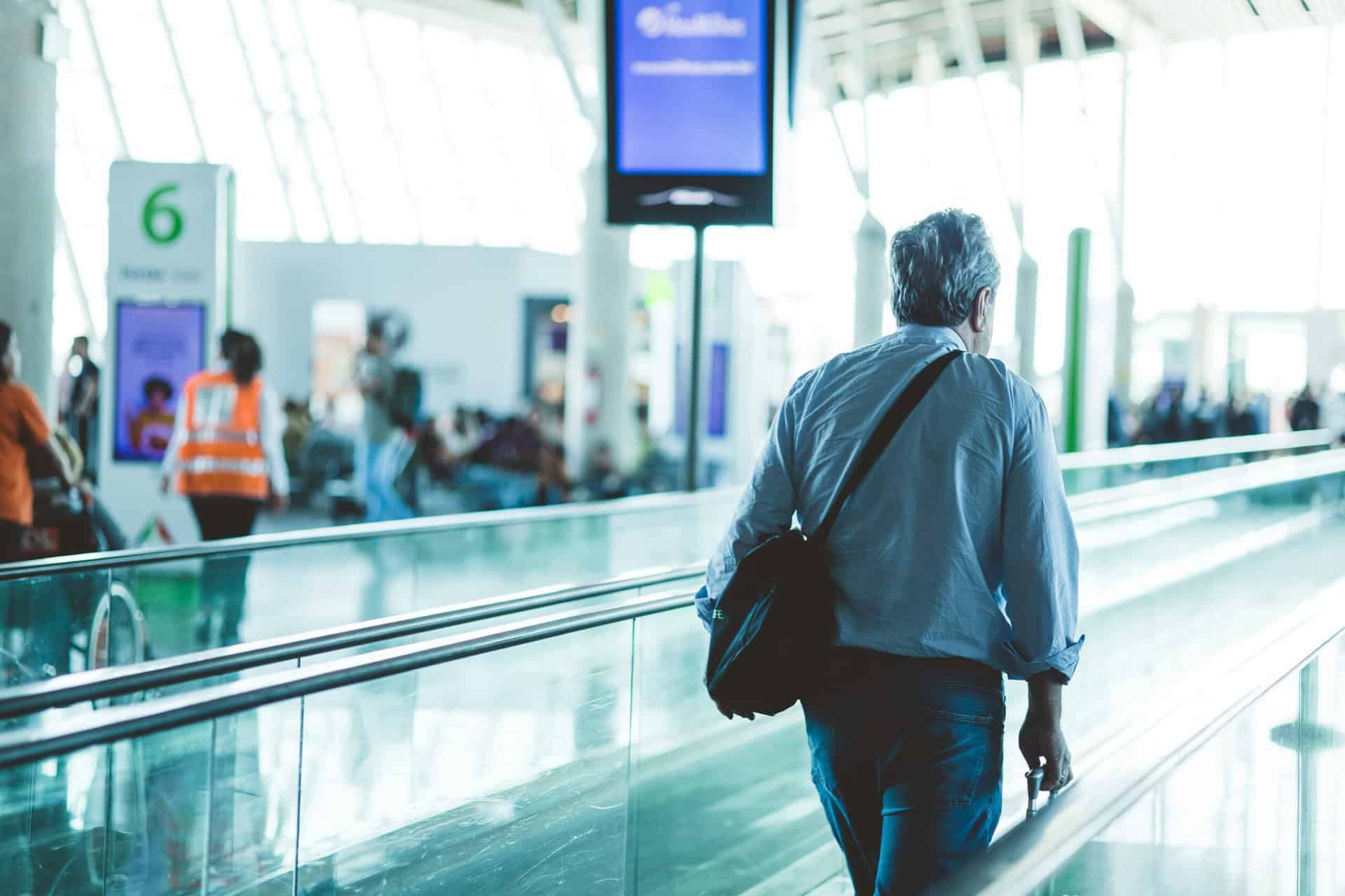 A traveller with a shoulder bag walking along an airport concourse beside a glass rail